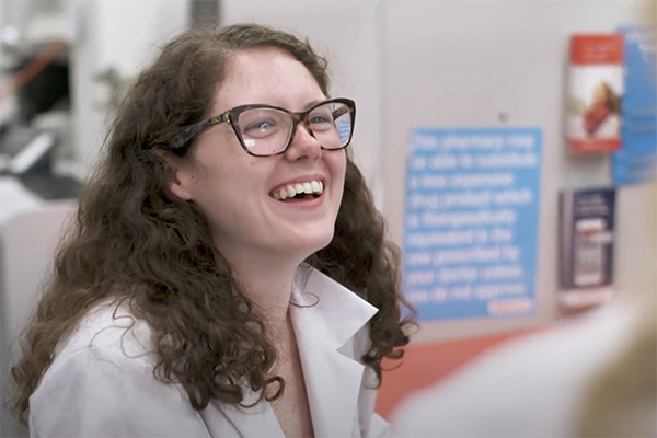 A woman with curly hair and glasses is smiling brightly, wearing a white lab coat. She appears to be in a professional or medical setting, with a blurred background that includes a blue sign and a red information flyer.