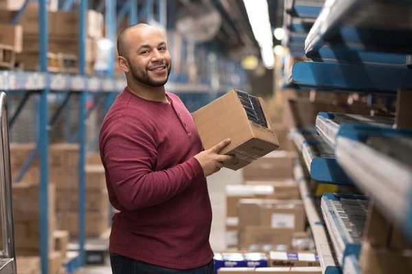 A man in a red long-sleeve shirt is holding a cardboard box while smiling at the camera. He is standing in a warehouse aisle surrounded by shelves stocked with various packages and items. The background shows more shelves and a lit pathway.