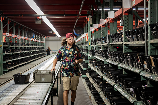 A person wearing a floral shirt, shorts, and a backward cap walks through a warehouse aisle lined with metal shelves and black bins. They are pushing a grey crate on a rolling conveyor belt with additional bins on it. The facility is brightly lit.