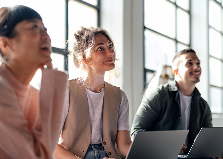 three people look up to someone speaking with their laptops in front of them