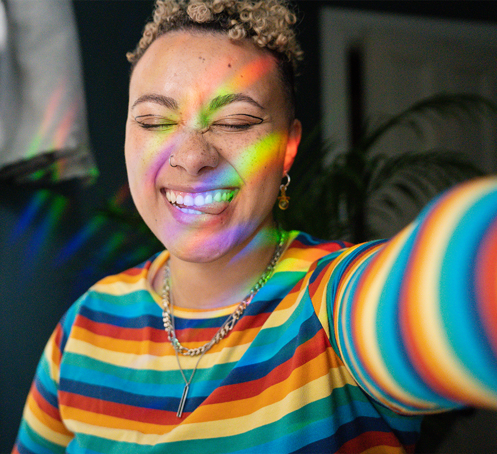 a woman poses for the camera with her eyes closed and tongue out, wearing a rainbow striped shirt and with the multicolor light displayed across her face