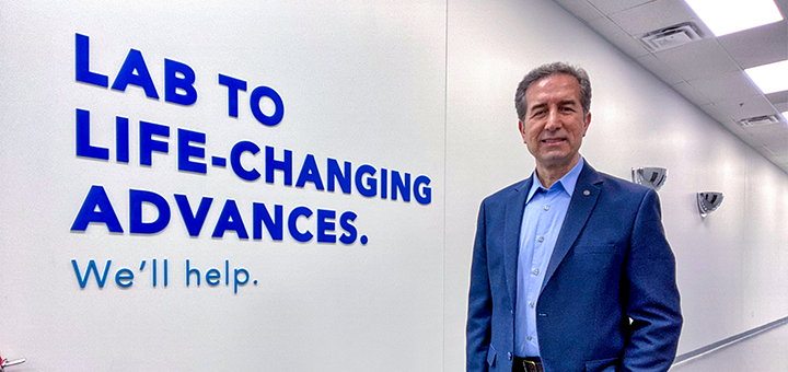 man with grey hair and a blue suit standing in a white hallway, in front of a sign that reads "Lab to life-changing advances. We'll help."