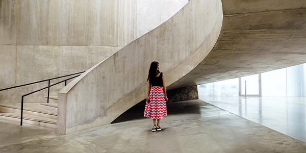 a woman in a black shirt and red and white striped skirt looking to her left, while standing in a monotone beige stone room with a curving staircase to her right