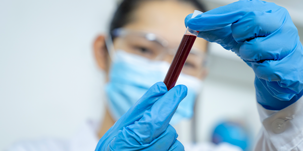 a scientist looking at a test tube full of red liquid. The scientist is out of focus, while her gloved hands and the test tube are in focus in the foreground.