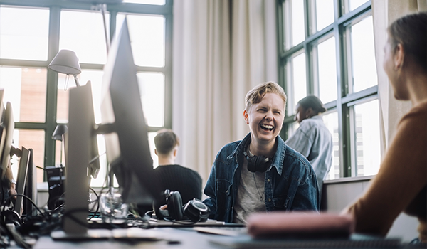 a person in an office sits in front of their computer, laughing while discussing something with the person to their left