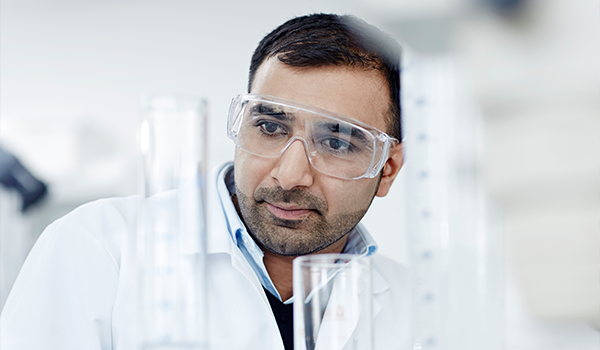 a male scientist with safety glasses looks at a large beaker in a lab