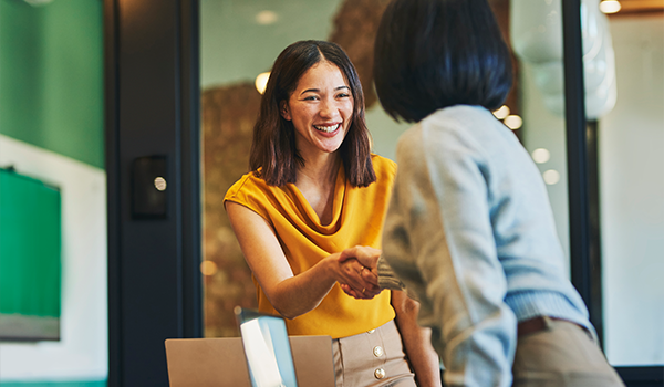 Woman in professional clothes shaking hands with another woman facing away from the camera in a brightly colored office