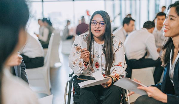 a woman sitting in a wheelchair leading a discussion in a group of people with other groups in the background