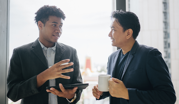 Two men standing side by side in suit jackets having a discussion in front of a window; the man on the left holds a tablet and the man on the right is holding a coffee cup