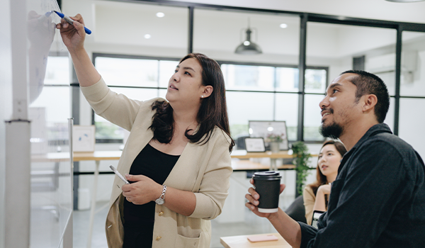 a woman writing on a whiteboard in an office while two co-workers look on