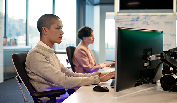 a person typing at a computer at their desk with another person at the adjacent workstation in the background