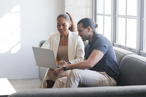 A LatinX male and multiracial female work together on a laptop while wearing smart casual clothing.