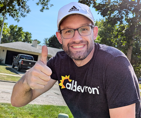 a man in a t-shirt with the Aldevron logo across the front giving a thumbs up standing outside in front of a road