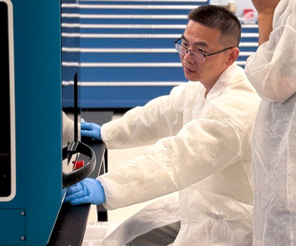a man in a lab, sitting down and working at a table with a large machine