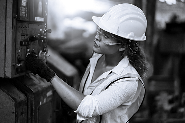 woman in safety helmet and glasses examines a machine in a factory