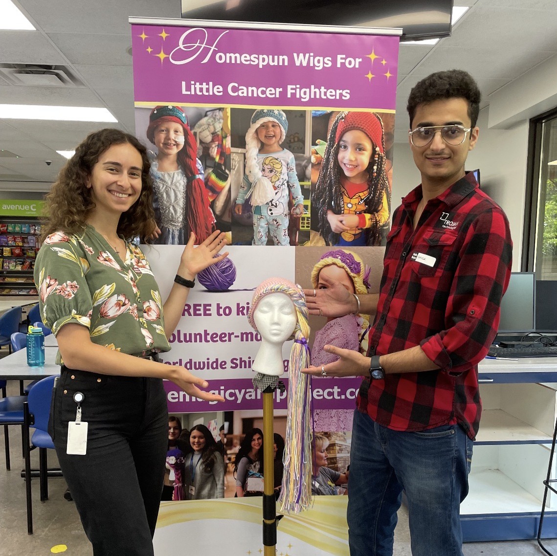 Two Trojan Technologies employees pose in front of a sign reading "Homespun Wigs for Little Cancer Fighters" and point to a wig knitted during their volunteer event