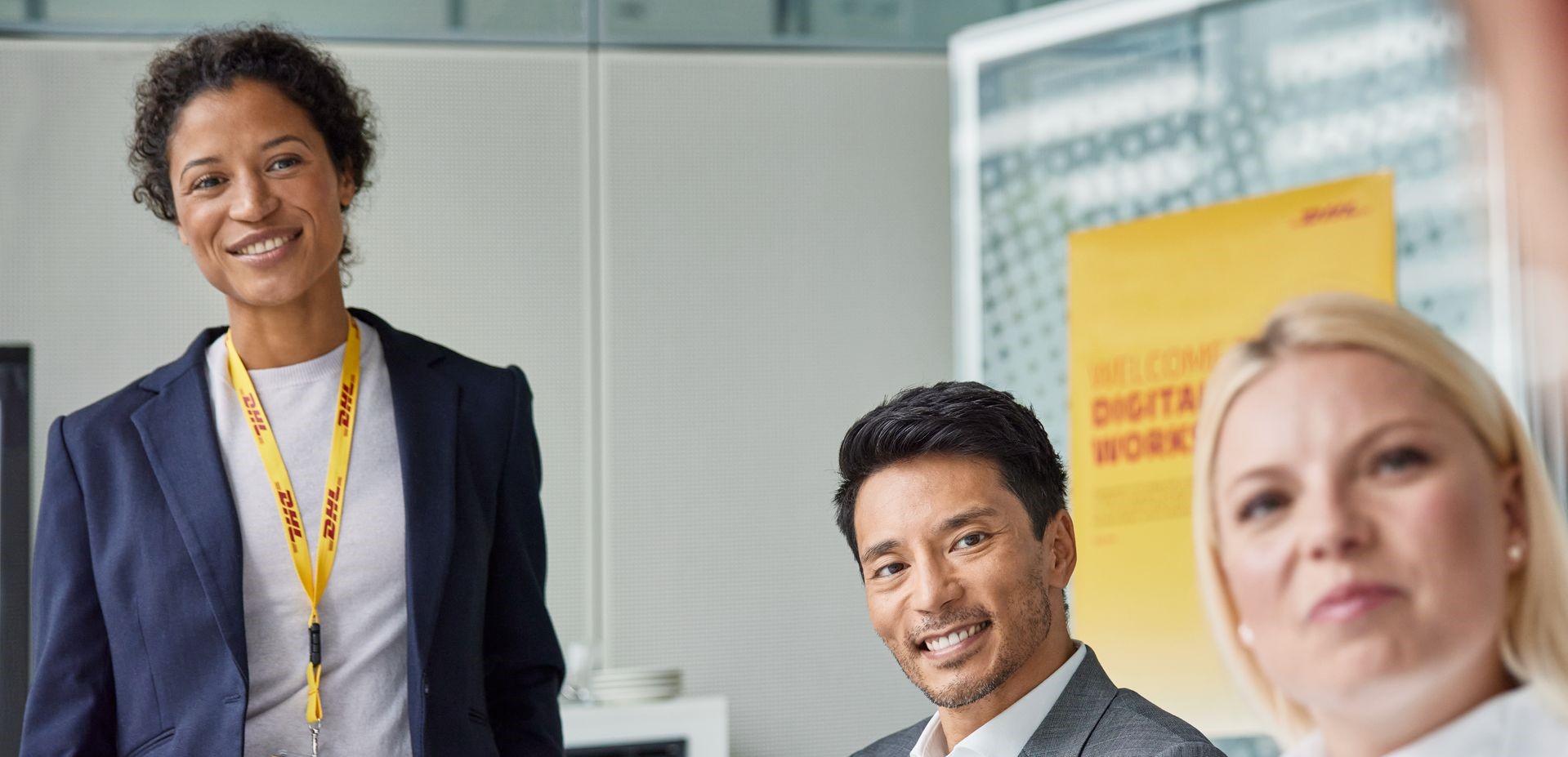 A diverse group of professionals in a bright office setting. A smiling woman wearing a navy blazer and a DHL-branded lanyard stands confidently, engaging with the team. Two seated colleagues, a man in a gray suit and a blonde woman, listen attentively. A yellow poster with 'Welcome Digital Workers' is visible in the background, adding to the professional and collaborative atmosphere.