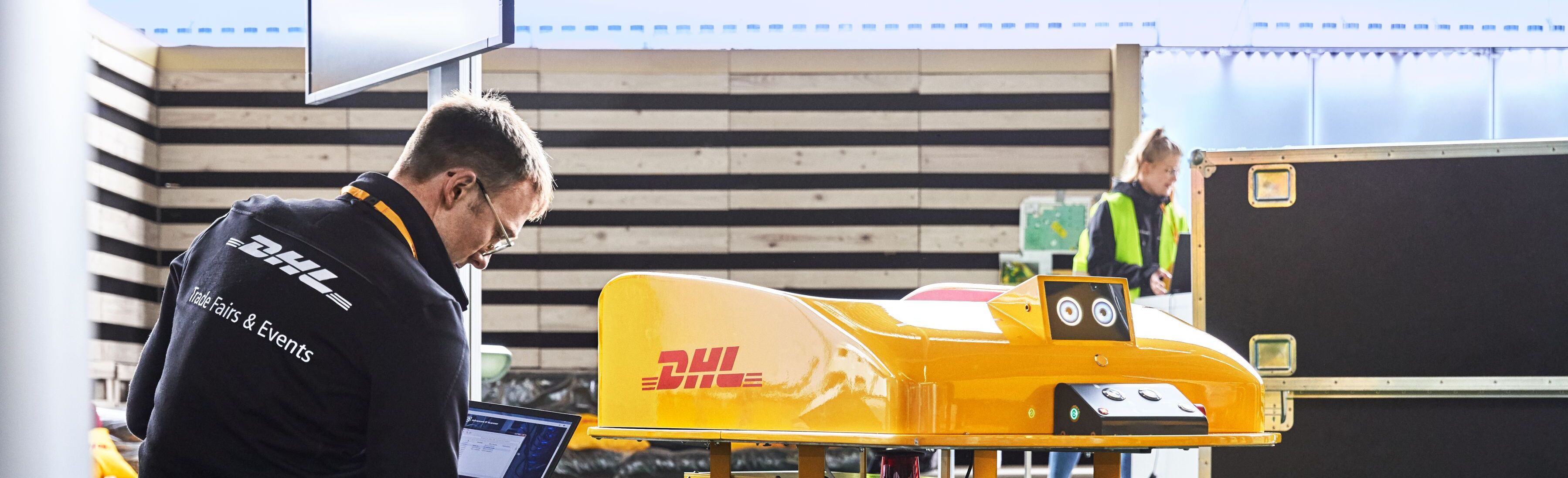 A man with a DHL Trade Fairs and Events jacket is looking at his laptop, while in front of him is a DHL warehouse robot