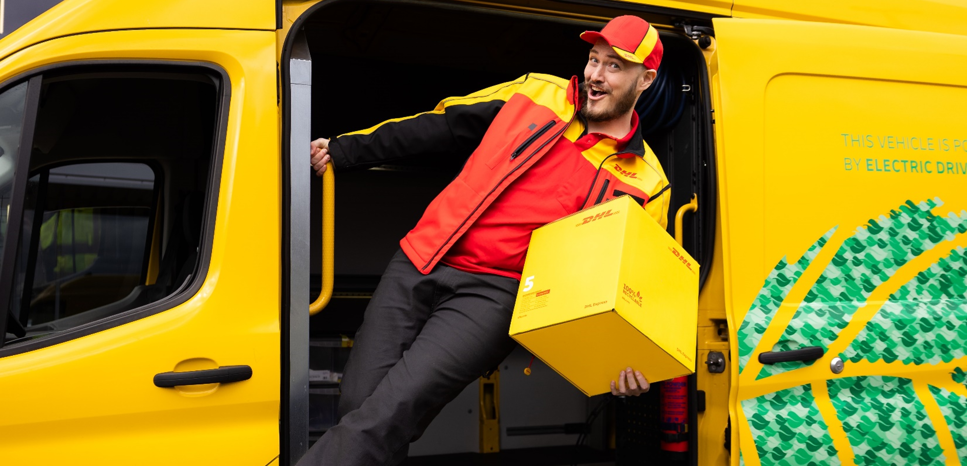 The image shows SirActionSlacks, the beloved DOTA2 content creator, dressed as a cheerful delivery driver in a DHL uniform, consisting of a red and yellow cap, red polo shirt, and red and yellow jacket. He is playfully leaning out of the open side door of a yellow DHL delivery van while holding a yellow DHL package in one hand. The van is labeled as being powered by electric drive, highlighting its eco-friendly features. SirActionSlacks is smiling energetically, adding his signature enthusiasm and charm to the scene.