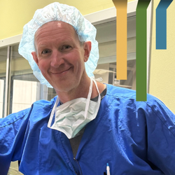 Male CRNA team member smiles in a scrub cap and blue scrubs.
