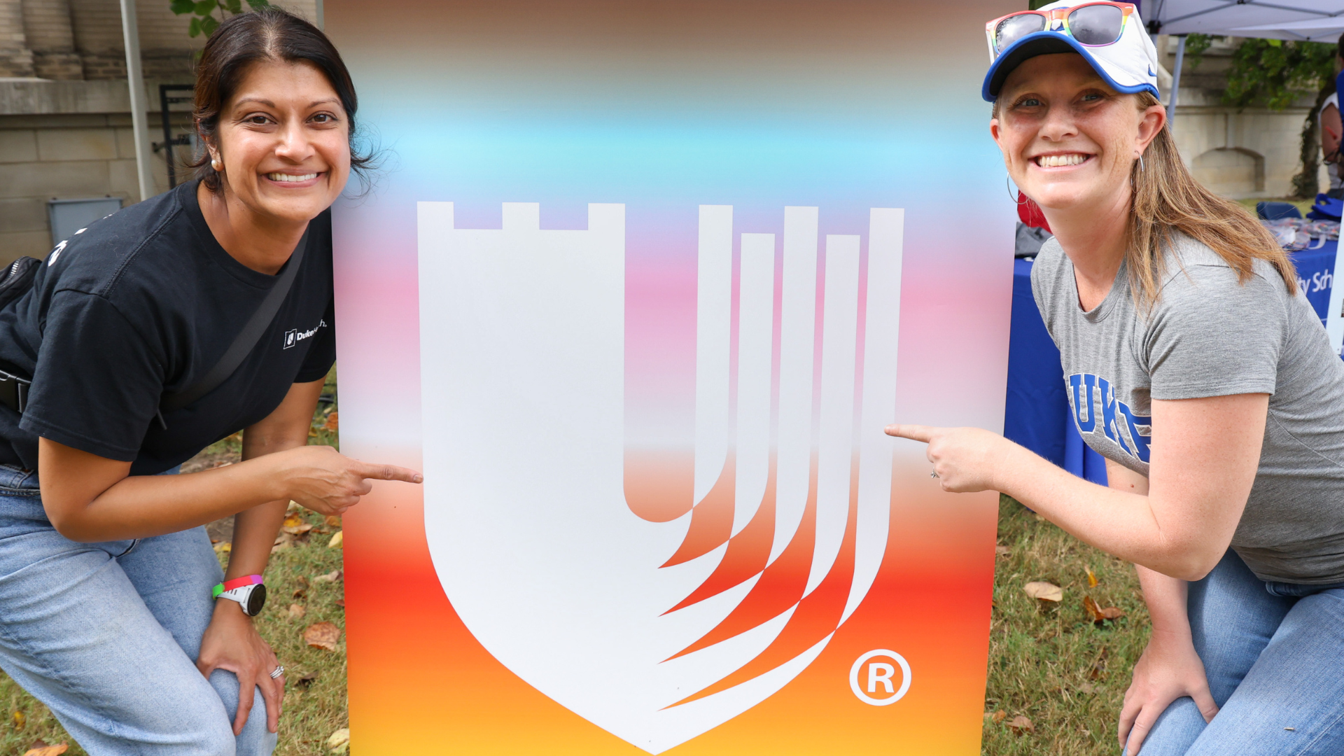 Two female team members smile and point to a colorful Duke shield.