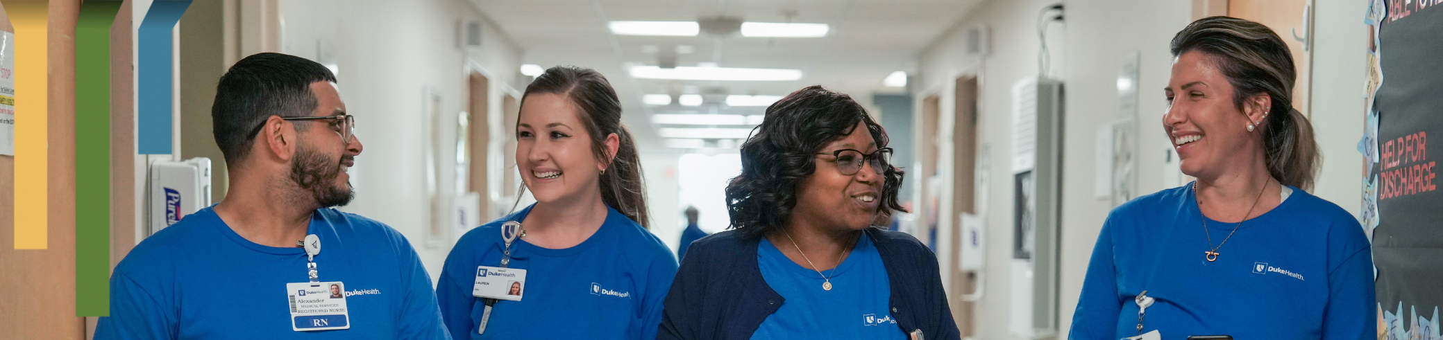 Four team members in Duke Health blue t-shirts smile and chat in a hallway.