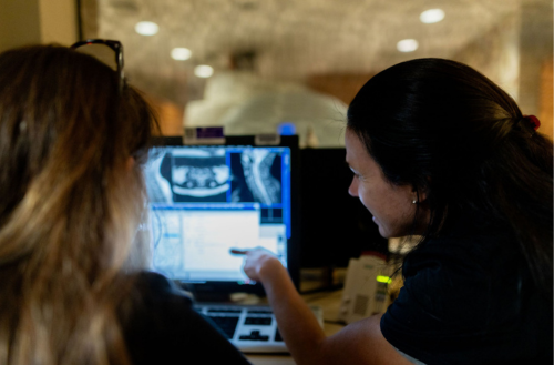 Two women talk over images on a computer screen with a CT scanner in the background.