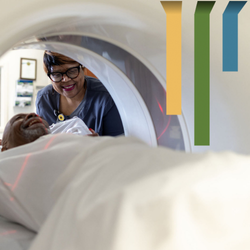 Female team member in blue scrubs and glasses helps a male patient into an imaging machine.
