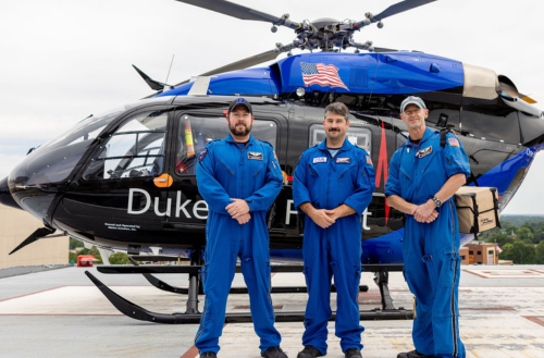Three male team members in life flight suits smile in front of a helicopter.