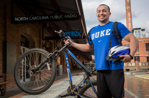 Male team member smiles in a Duke shirt on campus holding a bike.