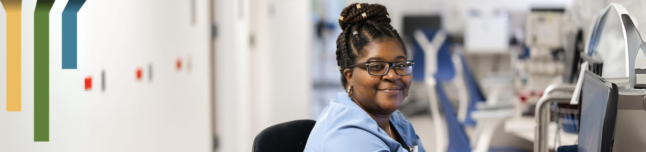 female nursing assistant smiles at a computer in blue scrubs.