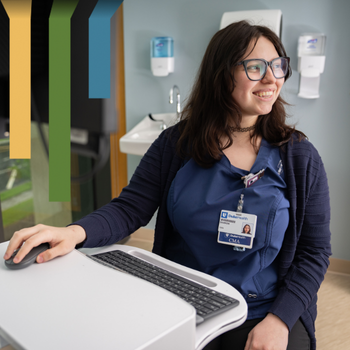 Female CMA in glasses and blue scrubs smiles at a patient while at a computer.