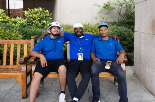 Team members sit on a bench in Duke blue shirts smiling together.