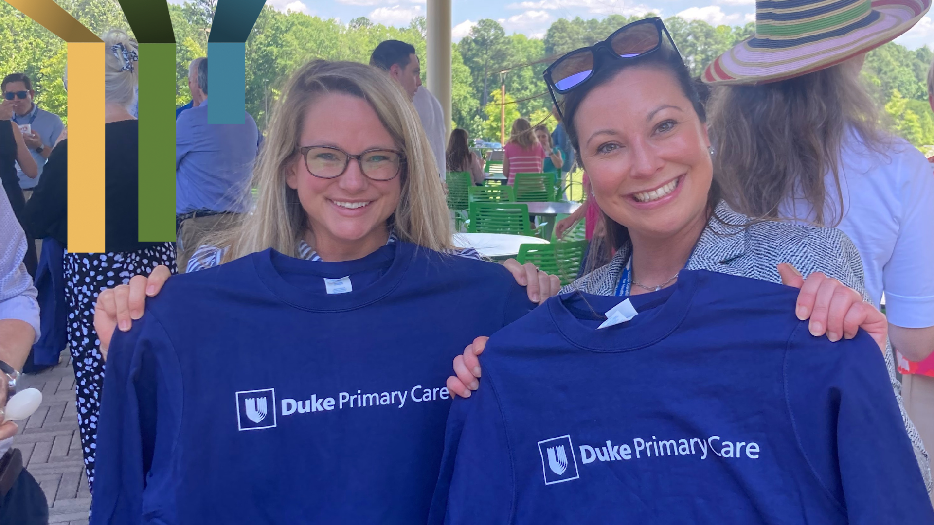 Two women smile while holding up two blue Duke Primary Care t-shirts.