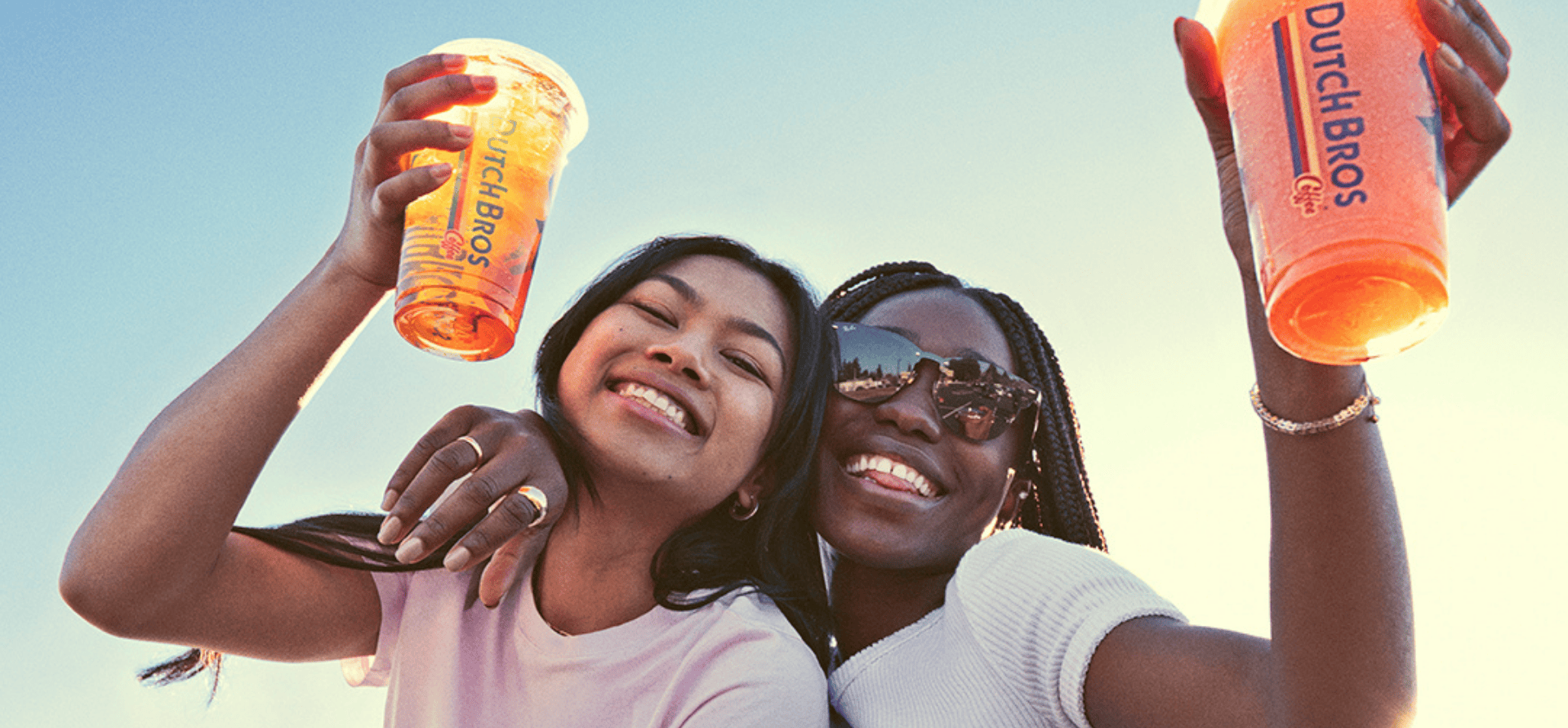 Two girls holding dutch bros drinks, smiling at sunset