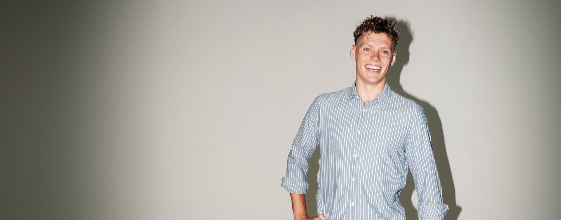 Junger Mann in gestreiftem Hemd steht lächelnd vor einer weißen Wand.