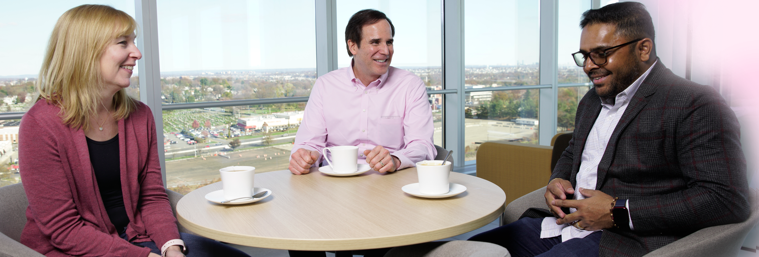 Three employees sitting in a common area with coffee mugs on the table while having a discussion 