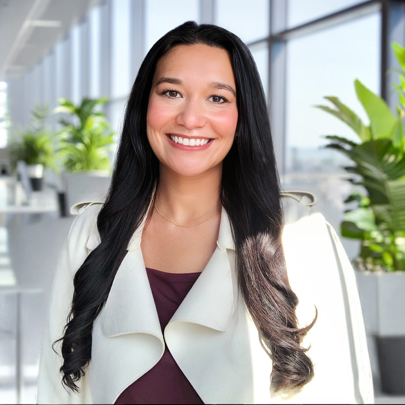 Professional headshot of a smiling woman with long dark hair, wearing a maroon top and cream jacket, in a bright office.