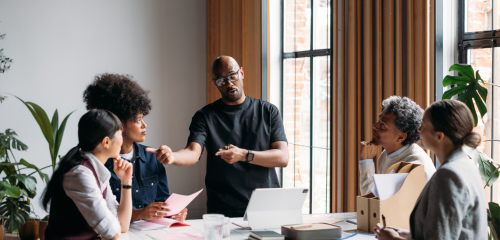 A diverse group of five colleagues collaborates in a bright, modern meeting room filled with plants.