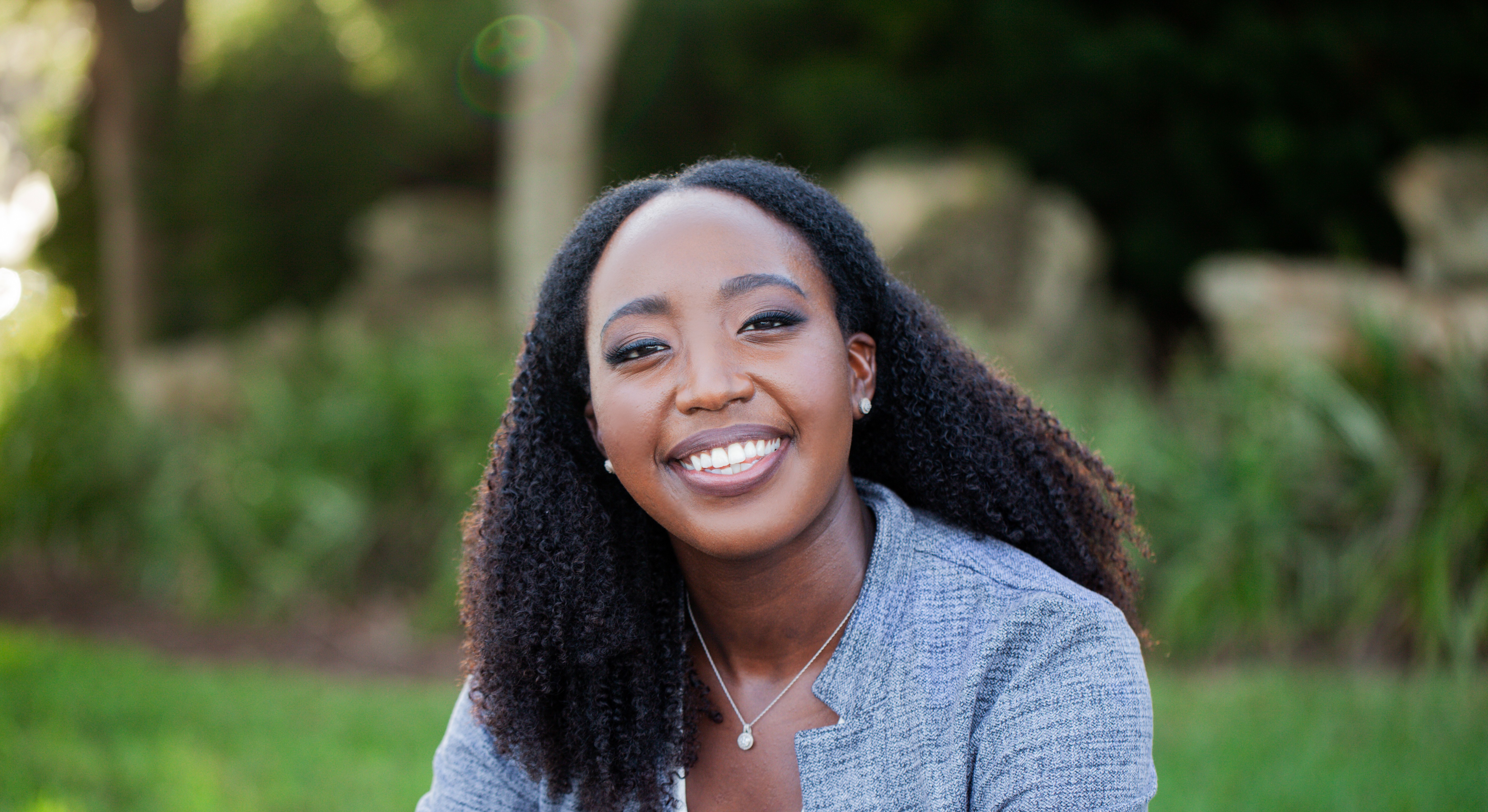    A smiling woman with long, curly black hair wears a gray textured blazer and delicate jewelry, including small stud earrings and a pendant necklace. The photo shows her from the shoulders up against a softly blurred green foliage background.