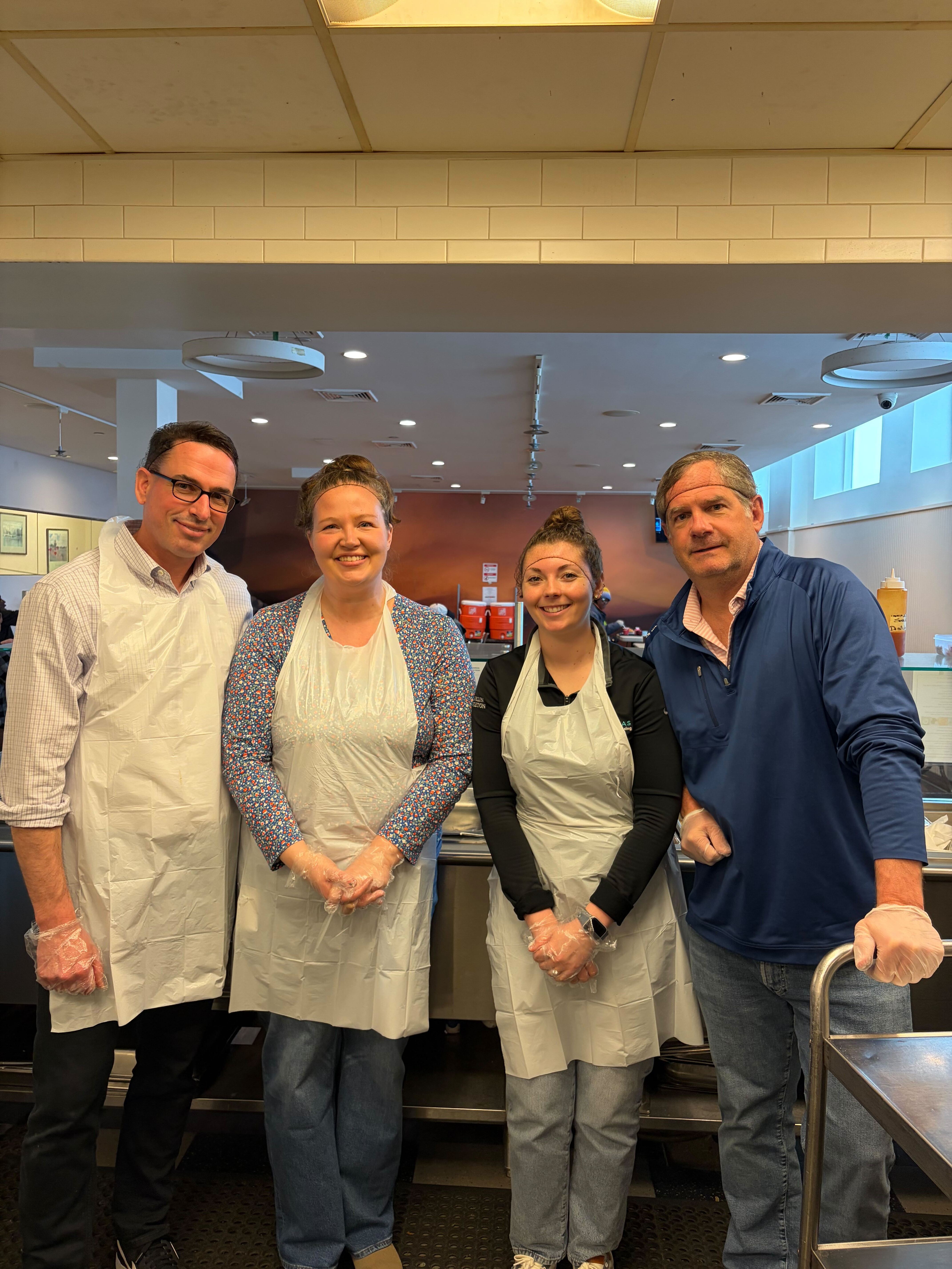Two men and two women stand smiling in a kitchen. Three wear white plastic aprons, and all wear disposable gloves.