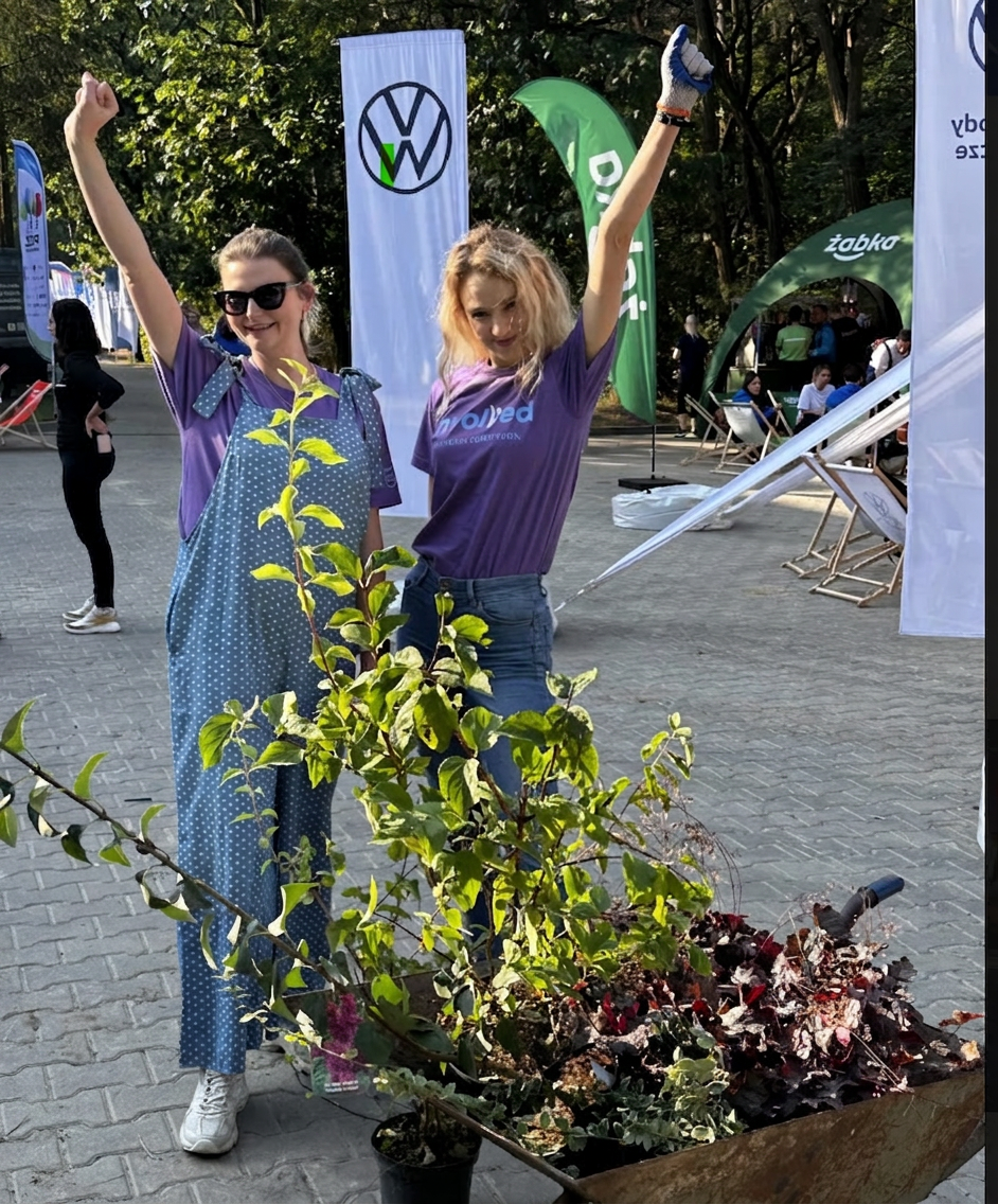 Two women in matching purple shirts celebrate outdoors beside a wheelbarrow filled with plants and a young tree—one in sunglasses and blue polka-dot overalls, the other blonde, wearing jeans and a gardening glove, with trees in the background.