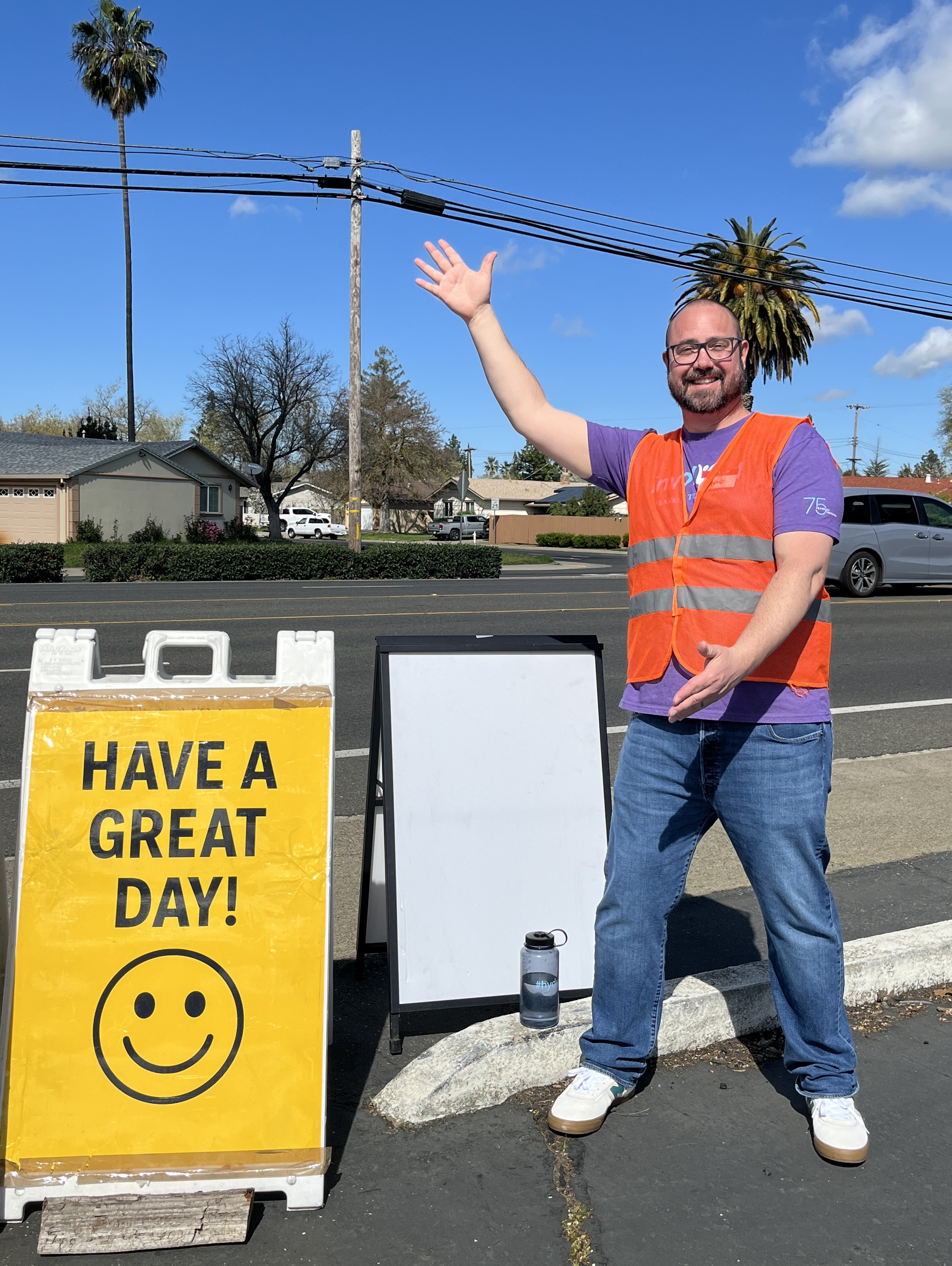 A man in an orange safety vest over a purple shirt stands by a road, smiling and waving next to a yellow A-frame sign that says "HAVE A GREAT DAY!" with a large smiley face below the text.