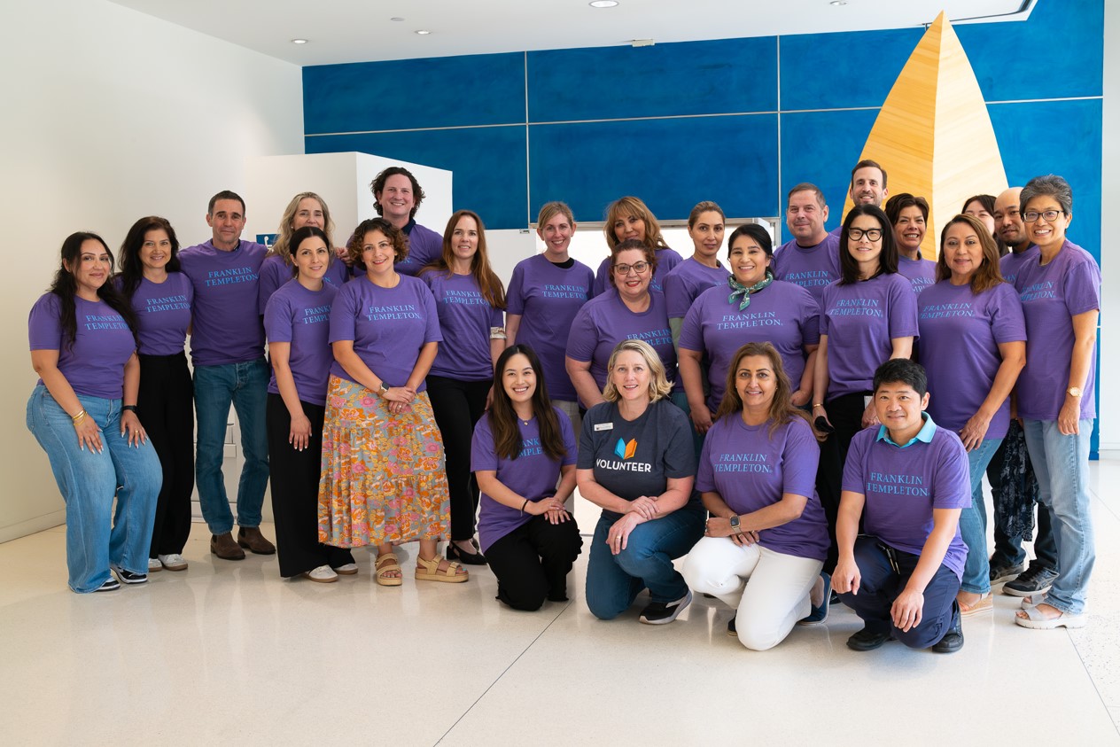 A group of  people pose indoors, most wearing matching purple "Franklin Templeton" t-shirts. They are arranged in standing back rows and a kneeling front row,.