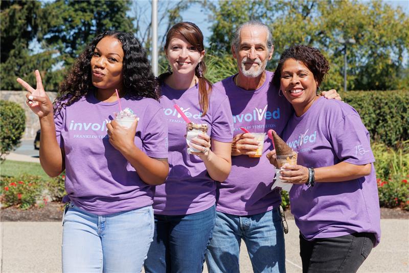 Four people wearing matching purple t-shirts with "involved" printed on them stand outdoors, smiling and holding frozen dessert cups. The person on the far left holds an ice cream cup and makes a peace sign. They stand in front of green trees under a sunny sky.