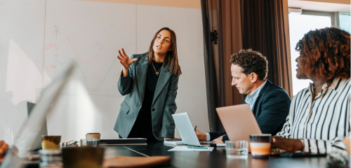 A woman in a grey blazer leads a business meeting, standing and gesturing in front of a whiteboard while her diverse colleagues listen attentively at a conference table.