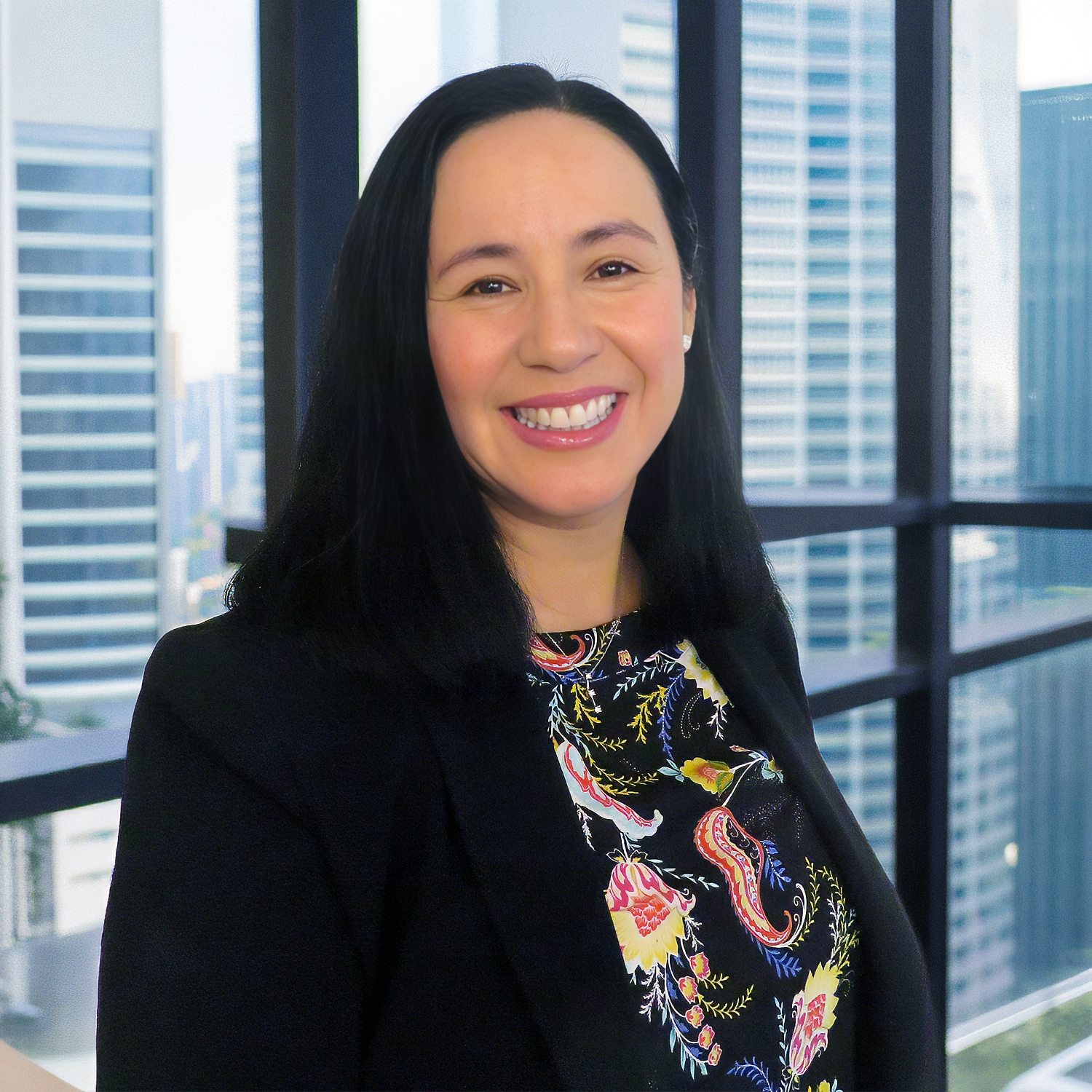 Smiling woman with long dark hair, wearing a black blazer and colorful patterned top, stands by a window with a city skyline in the background.