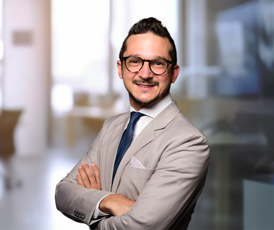 A smiling man with dark styled hair, stubble, and black-rimmed glasses stands with arms crossed. He wears a light gray business suit, white shirt, dark blue tie, and white pocket square. 