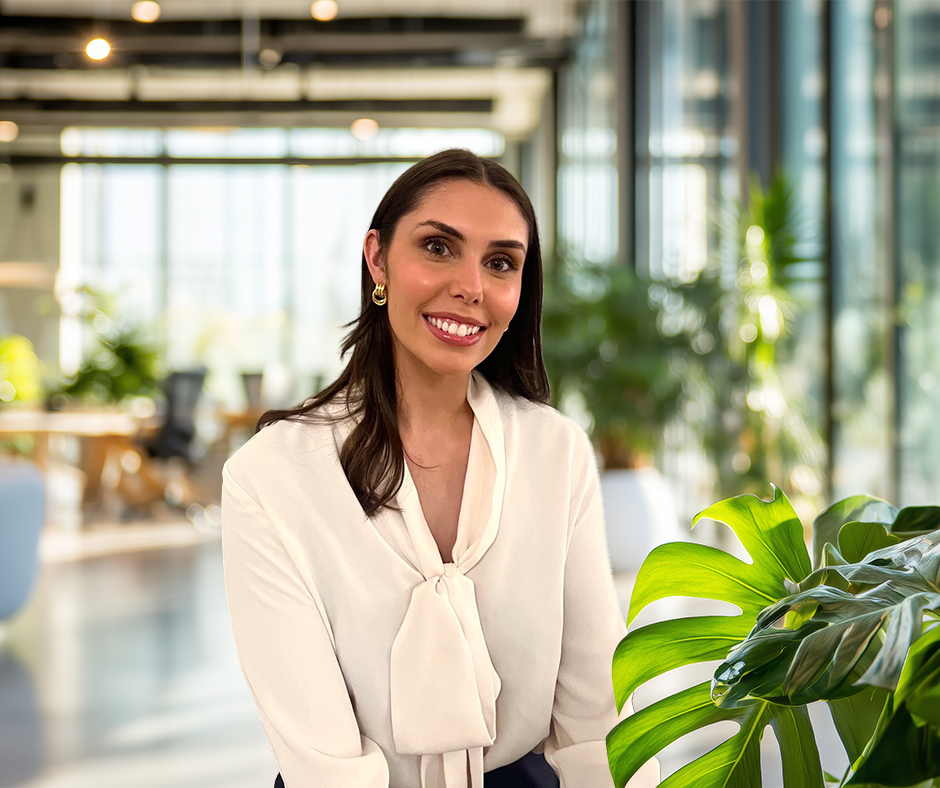 A smiling woman with long dark hair wearing a white blouse with a bow at the neckline, seated in a modern office with large windows and green plants, including a monstera leaf in the foreground.