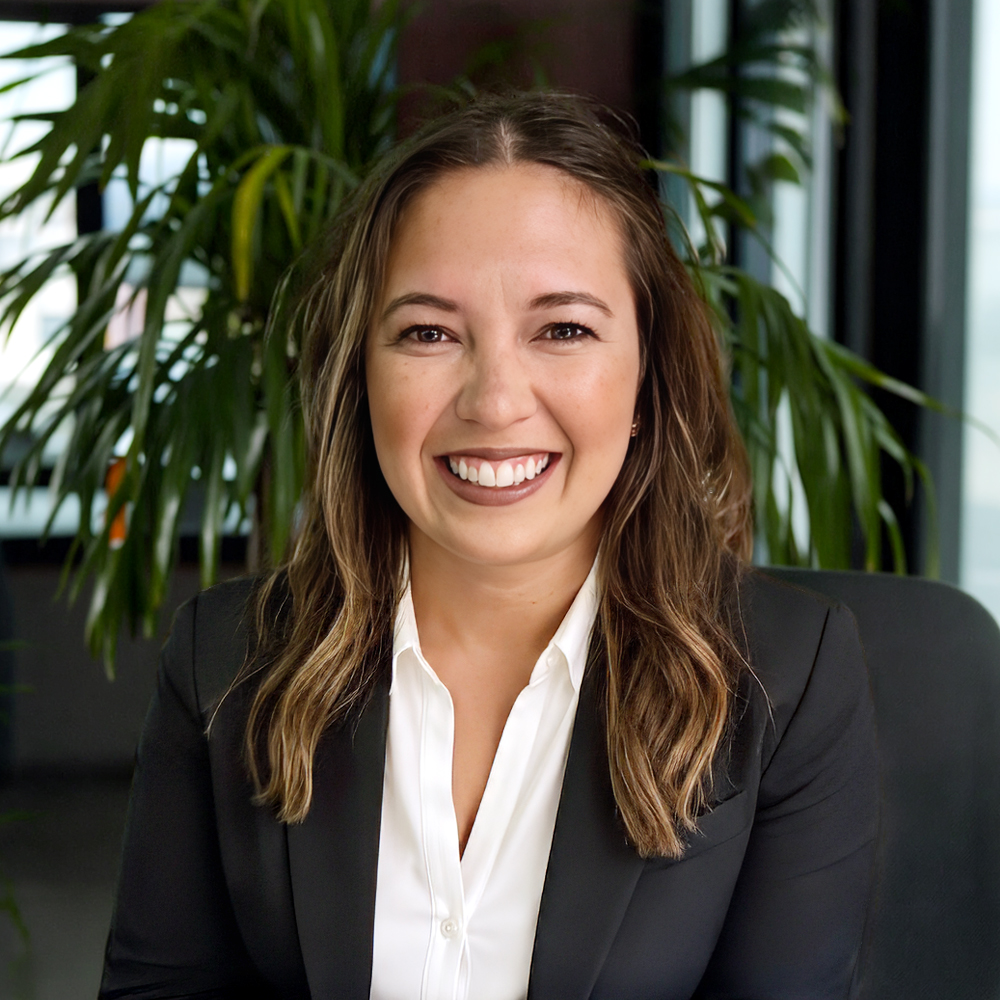 Professional headshot of a smiling woman with long wavy brown hair, wearing a white collared shirt and black blazer, with a large plant in the background.
